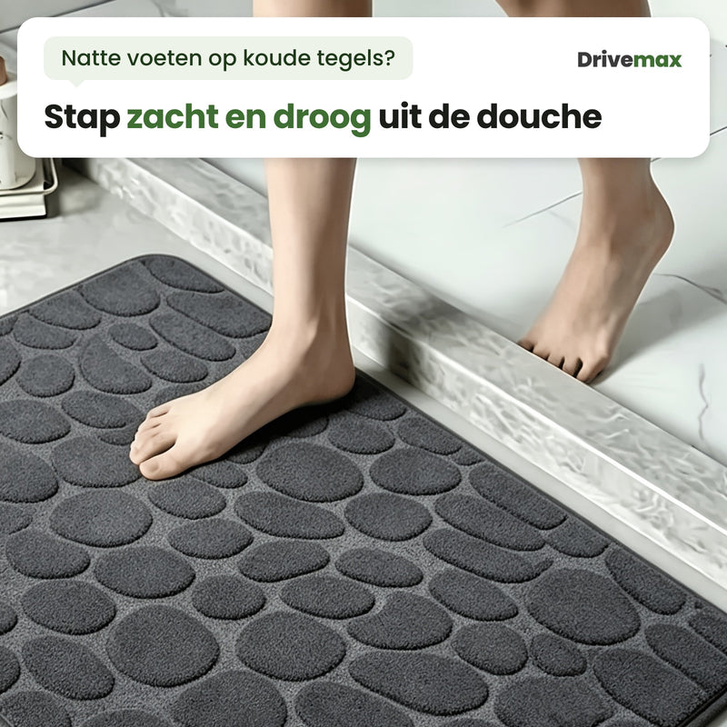 Iemand stapt uit de douche met een blote voet op de zachte kiezels van de donkergrijze Kiezova badmat, de andere voet nog op het opstapje: stap zacht en droog uit de douche